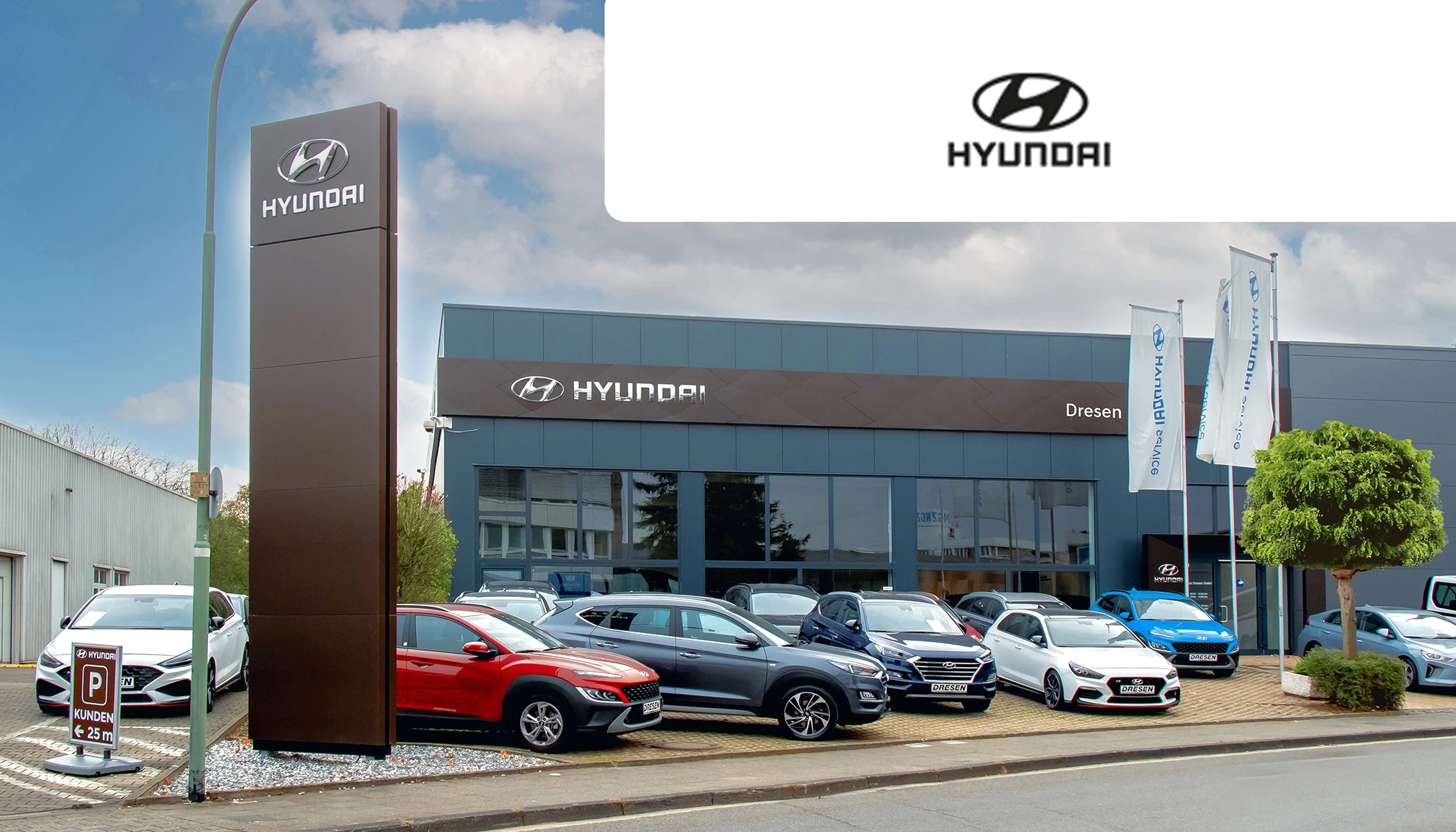 Hyundai Autohaus Dresen Standort mit Ausstellungsfahrzeugen auf dem Außengelände, hoher Hyundai-Steele, Flaggen und Glasfassade an einer Straße.